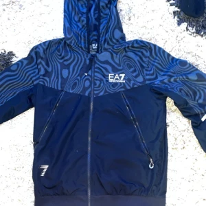 Mörkblå träningsbyxor och tröja från EA7 Emporio Armani - Snygga mörkblå träningsbyxor och tröja från EA7 Emporio Armani med unikt mönstrade detaljer längs sidorna.