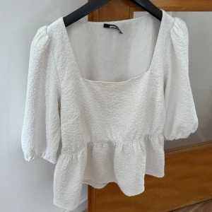 Vit blus med puffärmar från Gina Tricot - Säljer en söt vit blus från Gina Tricot med puffiga trekvartsärmar och volang nedtill. Blusen har en fyrkantig ringning och är tillverkad i ett strukturerat tyg som ger en trendig look.