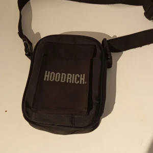 Svart axelväska från Hoodrich - Säljer en svart axelväska från Hoodrich med justerbart band och tydlig logga på framsidan. Väskan har en fyrkantig form och praktiskt fack framtill. Perfekt för att bära med sig det viktigaste.