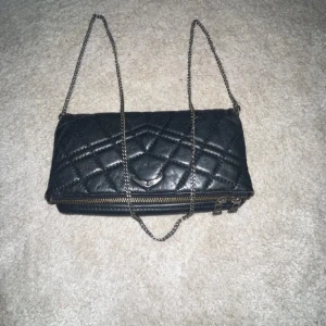 Svart quiltad axelväska med kedja - Snygg svart axelväska i quiltat skinnmönster med silverfärgad kedjerem och dragkedja. Insidan är fodrad och väskan stängs med dragkedja. Änvänd ett fåtal gånger💕