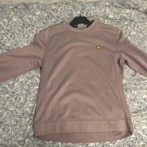 Ljuslila sweatshirt från Lyle & Scott - Säljer en stilren ljuslila sweatshirt från Lyle & Scott med klassisk rund halsringning och den ikoniska gula örnloggan på bröstet. Tröjan har långa ärmar och är tillverkad i mjukt material som passar perfekt till vardags.