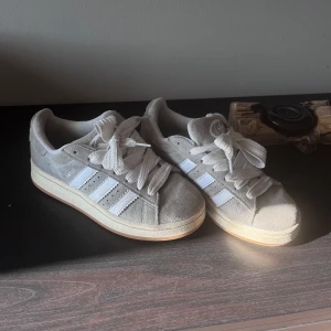 Grå Adidas Campus sneakers med vita detaljer - Säljer ett par grå Adidas Campus sneakers med vita ränder och vit sula. Skorna har breda vita skosnören och är i mocka med klassisk Adidas-logga på plösen. Perfekta för dig som gillar en stilren och tidlös look. Använda några få gånger på crossbanan därav lite dammigt på mockan. Original skolådan finns med och skickas med!