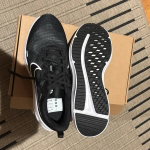 Nike Downshifter 12 svart/vit sneakers - Säljer ett par helt nya svarta Nike Downshifter 12 sneakers med vit sula. Storlek: 40,5. Skorna har snörning och en sportig design, perfekta för träning eller vardagsstil. Ovandelen är i syntetmaterial och mesh för bra andningsförmåga. Köpta för 800kr, mitt pris 400kr, vid frågor kontakta gärna.