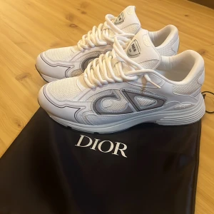 Vita Dior B30 sneakers - Säljer ett par vita Dior B30 skor med reflekterande CD-logga på sidan. Skorna har snörning, rund tå och platt sula. Riktigt bekväma och passar perfekt till våren och sommaren. Använd ett fåtal gånger. Priset är ej hugget i sten så var inte rädda att ställa frågor. Kan även tvätta skorna innan jag fraktar dem😊