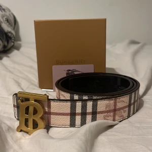 Rutig Burbery bälte med guldfärgat spänne - 115 cm. Snyggt Burbery bälte med rutigt mönster i beige, svart och rött. Bältet har ett stort guldfärgat 'TB'-spänne och är gjort i skinn. Perfekt accessoar för att maxa din stil. Nästan helt oanvänd och helt ren. PRISET KAN DISKUTERAS.
