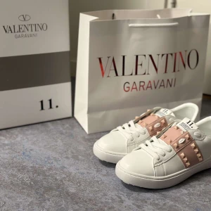Valentino Garavani sneakers med nitar - Säljer ett par vita Valentino Garavani sneakers med breda ljusrosa band över vristen dekorerade med guldiga nitar. Skorna har rund tå, platt sula och klassisk snörning. Kommer med originalkartong och påse.