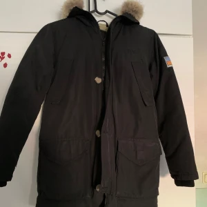 Svart parkajacka med fuskpäls från Svea - Svart parkajacka från Svea med huva och fuskpäls. Jackan har dragkedja, knappar och flera stora fickor framtill samt patch på ärmen. Fodrad insida i beige för extra värme.