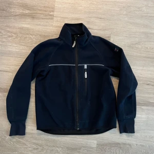 Svart fleecejacka från Polarn O. Pyret - Mörkblå fleecejacka från Polarn O. Pyret med hög krage och hel dragkedja framtill. Jackan har en bröstficka med dragkedja och en tunn vit linje över bröstet. Perfekt för kyliga dagar.