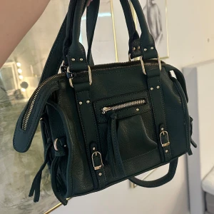 Mörkgrön handväska med gulddetaljer - Snygg mörkgrön handväska med guldfärgade spännen och dragkedjor. Väskan har både handtag och axelrem, samt en praktisk ytterficka med dragkedja och dekorativa spännen framtill. Invändigt randigt foder och flera fack för bra organisering. Köpt i Spanien 