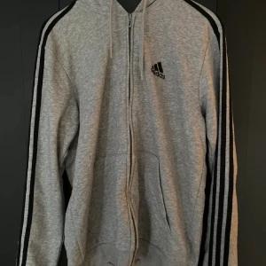 Grå adidas zip up - Säljer en grå hoodie från Adidas med dragkedja. Klassisk Adidas-logga på bröstet och huva med dragsko. Perfekt för en sportig och avslappnad stil.