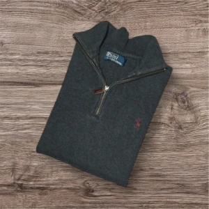 Mörkgrå halvzip tröja från Polo Ralph Lauren - Mörkgrå långärmad tröja från Polo Ralph Lauren med halv dragkedja och liten röd broderad logga på bröstet. Tröjan har hög krage och är tillverkad i mjukt material, perfekt för kyligare dagar.