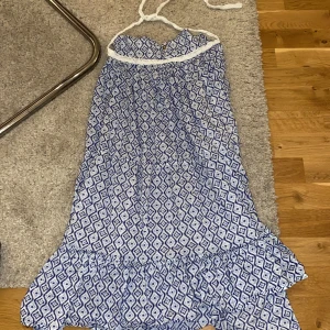 Blåvit mönstrad halterneckklänning med volang - Säljer en blåvit halterneckklänning med geometriskt mönster och volang nertill. Klänningen har virkade detaljer vid halsen och knytning i nacken. Perfekt för varma dagar och har en luftig passform.
