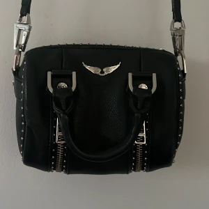 Svart handväska med silverdetaljer från Zadig & Voltaire - Cool svart handväska från Zadig & Voltaire med silverfärgade metalldetaljer, nitar längs kanterna och dubbla dragkedjor framtill. Väskan har både handtag och lång axelrem, samt en ikonisk vinglogga i metall på framsidan. Köparen står även för frakt!