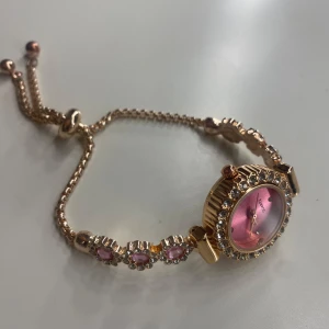 Guldig klocka med rosa urtavla och stenar - Säljer en elegant klocka med guldig länk och rosa urtavla. Armbandet är dekorerat med rosa stenar och gnistrande detaljer runt urtavlan. Perfekt accessoar för dig som gillar bling och färg. Super skön att ha på sig och passar fint med mycket🥰 den är helt ny har ej använt den 😁priset går att diskuteras💞den är justerbar så den passar💞
