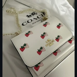 Vit axelväska med jordgubbsmönster från Coach - Superfin vit axelväska från Coach med kedjerem och sött jordgubbsmönster i rött och rosa. Väskan har guldfärgade detaljer och logga framtill. Perfekt för dig som vill ha något unikt och lekfullt i din accessoargarderob!