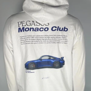 Vit hoodie med tryck från Pegasus - Säljer en vit hoodie från Pegasus med tryck på både fram- och baksidan. Framsidan har en liten blå logotyp och text, medan baksidan pryds av en stor bild av en blå sportbil och texten 'Monaco Club'. Hoodien har en klassisk design med dragsko i huvan och en stor ficka framtill.