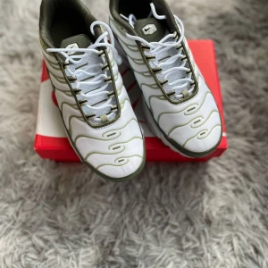 Nike Air Max Plus Tn sneakers i vitt och olivgrönt - Nike Air Max Plus Tn sneakers i vitt med olivgröna detaljer och sula. Skorna har snörning, rund tå och den klassiska Tn-loggan på hälen. Perfekta för dig som gillar en sportig och modern stil.
