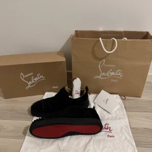 Svarta sneakers med nitar från Christian Louboutin - Säljer ett par svarta Christian Louboutin Louis Junior Spikes Flat sneakers i mocka med svarta nitar på tån och klassisk röd sula. Skorna har snörning och en stilren, lyxig look. Kommer med originalkartong och dustbag. Kvitto fins och extra sko snören köpta i Danmark i butik