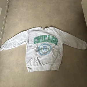 Vit sweatshirt med CHICAGO-tryck från FB Sister - Vit sweatshirt från FB Sister med grönt och blått CHICAGO-tryck på bröstet. Klassisk rund halsringning och ribbade muddar vid ärmslut och nederkant. Perfekt för en avslappnad stil.