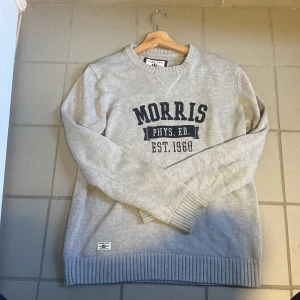 Grå tröja från Morris med tryck - Säljer en grå långärmad tröja från Morris med mörkblå texttryck på bröstet: 'Morris Phys. Ed. Est. 1968'. Tröjan har ribbade muddar vid ärmslut och nederkant samt rund halsringning.