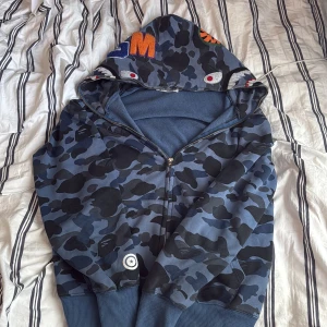 Bape hoodie mörkblå - Säljer en blå camo hoodie från A Bathing Ape med dragkedja och broderade detaljer på luvan. Hoodien har ribbade muddar och ett unikt camouflagemönster. Perfekt för dig som vill sticka ut med streetwear. Den är äkta. Står XL MEN passar M