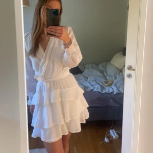 Vit omlottklänning med volanger - Säljer en superfin vit omlottklänning med volanger och knyt i midjan. Klänningen har långa ärmar och v-ringning, samt flera lager i kjolen som ger en luftig känsla. Perfekt för sommarens alla tillfällen! Klänningen är köpt från Nelly!🥰