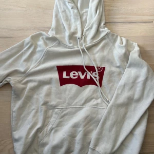 Vit hoodie från Levi's - Vit hoodie från Levi's med röd logga framtill. Klassisk modell med huva och känguruficka. Perfekt för en avslappnad stil och enkel att matcha med jeans eller leggings.