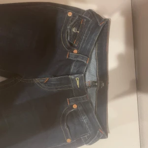 Mörkblå jeansbyxor från True Religion - Säljer ett par mörkblå jeansbyxor från True Religion med klassiska kontrastsömmar och dekorativa ficklock bak. Byxorna har låg midja och utsvängda . Snygga detaljer med logga på bakfickan och knappar i silverfärg.