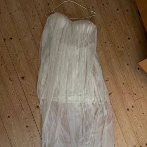 Vit långklänning från LACE & BEADS - Säljer en elegant vit långklänning från LACE & BEADS helt oanvänd. Klänningen är axelbandslös med en lätt transparent, plisserad tyll över hela plagget. Perfekt för speciella tillfällen när du vill känna dig extra fin.