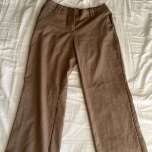 Bruna kostymbyxor från Monki - Säljer ett par bruna kostymbyxor från Monki med normal passform och raka ben. Byxorna har hällor för bälte och stängs med dragkedja och knapp. Perfekta för en stilren look.