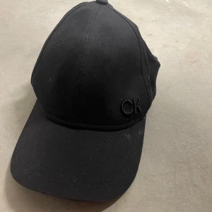Svart keps från Calvin Klein - Snygg svart keps från Calvin Klein med diskret CK-broderi framtill. Klassisk böjd skärm och justerbar passform. Tillverkad i bomull för en bekväm känsla och stilren look. Nypris 499kr