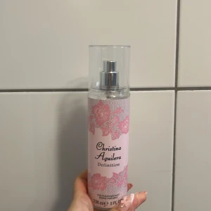 Christina Aguilera Definition Body Mist - En body mist från Christina Aguilera med namnet Definition. Flaskan är cylinderformad och genomskinlig med rosa etikett dekorerad med spets- och blommönster. Spraymunstycke och genomskinligt lock. Rymmer 236 ml.