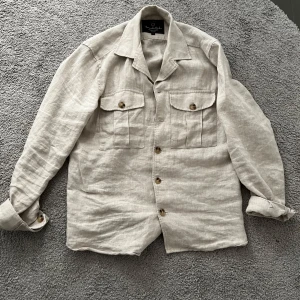 Beige linneskjorta från Bondelid - Snygg beige skjorta i linne från Bondelid. Skjortan har två stora bröstfickor med lock och knappar framtill. Perfekt för en avslappnad och stilren look. Klassisk krage och långa ärmar.