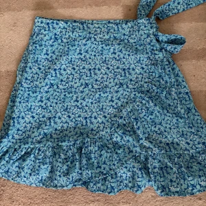 Blommig omlottkjol från FB Sister - Säljer en blå blommig omlottkjol från FB Sister i storlek M. Kjolen har volang nertill och knyts i midjan för en snygg passform. Perfekt för dig som gillar färgglada och mönstrade plagg!