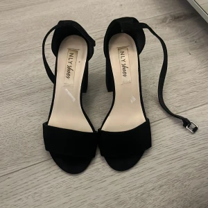 Svarta klackskor från NLY Shoes - Säljer ett par svarta klackskor från NLY Shoes med öppen tå och bred klack. Skorna har en enkel rem över tårna och justerbar ankelrem med spänne. Perfekta för dig som vill ha en stilren och klassisk look. Andvänt dem ett par gånger men nu på senaste har dem bara stått 