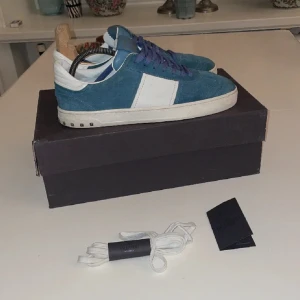 Blåa sneakers från Valentino - Säljer ett par blåa sneakers från Valentino med vit panel på sidan och vita detaljer. Skorna har snörning, rund tå och platt sula. Yttermaterialet är mocka och de levereras med extra vita skosnören.