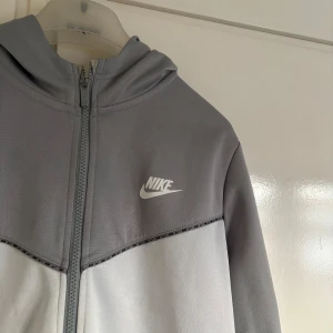 Grå zip-hoodie från Nike - Snygg tvåfärgad hoodie från Nike i ljusgrått och mörkare grått. Jackan har dragkedja hela vägen, huva och två fickor framtill. Perfekt för en sportig och avslappnad stil.