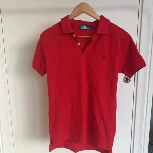 Röd pikétröja från Polo Ralph Lauren - Klassisk röd pikétröja från Polo Ralph Lauren med blå broderad logga på bröstet. Tröjan har korta ärmar, krage och knappar vid halsen. Perfekt för en stilren och sportig look.