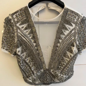 Vit och silverglittrig croppad topp med pärlor och paljetter - Säljer en supercool croppad topp i vitt med massor av silvriga pärlor, paljetter och glittriga detaljer. Djup v-ringning och korta ärmar. Perfekt för att sticka ut på festen!