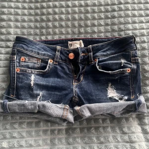 Lågmidjade Jeansshorts - Säljer ett par mörkblå jeansshorts med slitningar och upprullade benslut. Shortsen har klassiska fem fickor, kopparfärgade nitar och knapp, samt låg midja. Perfekta för sommaren! Midjemåttet är 33 men kan stretchas upp till 35 cm