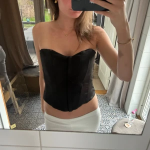 Svart korsett-topp - Säljer en svart axelbandslös korsett-topp med markerad midja och lätt glansigt material. Toppen har en stilren och elegant design som passar perfekt till både jeans och kjol.