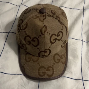 Beige keps från Gucci med GG-mönster - Snygg beige keps från Gucci med klassiskt brunt GG-mönster över hela kepsen. Kepsen har bruna detaljer och justerbart bälte bak i skinn. Perfekt accessoar för att lyfta din stil.