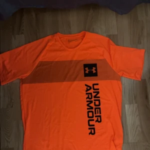 Orange t-shirt från Under Armour - Säljer en orange t-shirt från Under Armour med svart logga och text på framsidan. T-shirten har rund halsringning och är kortärmad, perfekt för träning eller sportiga aktiviteter.
