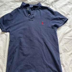 Blå pikétröja från Polo Ralph Lauren - Mörkblå pikétröja från Polo Ralph Lauren med klassisk krage och två knappar. Slim fit-modell med korta ärmar och röd broderad logga på bröstet. Perfekt för en stilren och sportig look.
