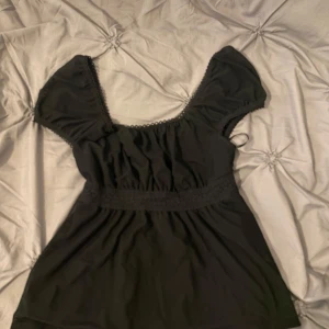 Svart topp med knytband och puffärmar - Säljer en svart topp med korta puffärmar och söt spetskant vid halsringningen. Toppen har knytband i midjan och är tillverkad i ett mjukt material. Perfekt för dig som gillar en feminin och enkel stil.