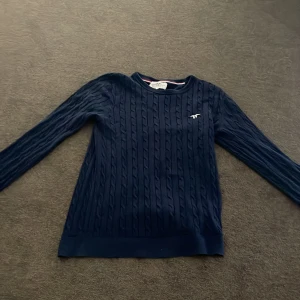 Mörkblå kabelstickad tröja från Hampton Republic st 170 - Säljer en mörkblå kabelstickad tröja från Hampton Republic med broderad logga på bröstet. Tröjan har rund halsringning och långa ärmar, perfekt för en stilren look. St 170