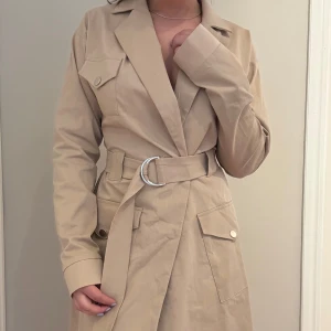 Beige trenchcoat med bälte från NAKD - Tunn beige trenchcoat med markerad midja och bälte med silverfärgat spänne. Jackan har stora fickor med knappar samt klassisk krage. Bara testat, aldrig använd. Storlek 36, säljer för 100kr 💕