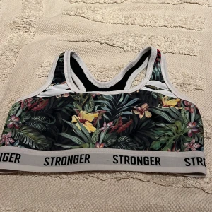 Blommig sporttopp från Stronger - Sporttopp från Stronger med tropiskt blommönster i grönt, gult, rött och rosa. Elastiskt band med logga, vita kanter och brottarrygg. Perfekt för träning och ger en snygg, sportig look.
