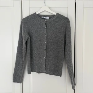 Grå kofta från Zara med pärlknappar - Mysig grå kofta från Zara med lång ärm och rund halsringning. Koftan är stickad och har snygga pärlknappar framtill som ger en stilren touch. Perfekt att slänga över en topp eller klänning för en chill look.