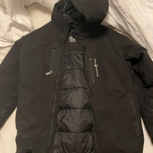 Svart Sail Racing jacka - Svart vinterjacka från Sail Racing med huva och dragkedjor på bröstet. Jackan har quiltad insida, logga på bröstet och ärmarna samt en stilren look. Perfekt för kalla och regniga dagar. Jackan är inköpt för ungefär 3.500. Modellen går ej att köpa längre 